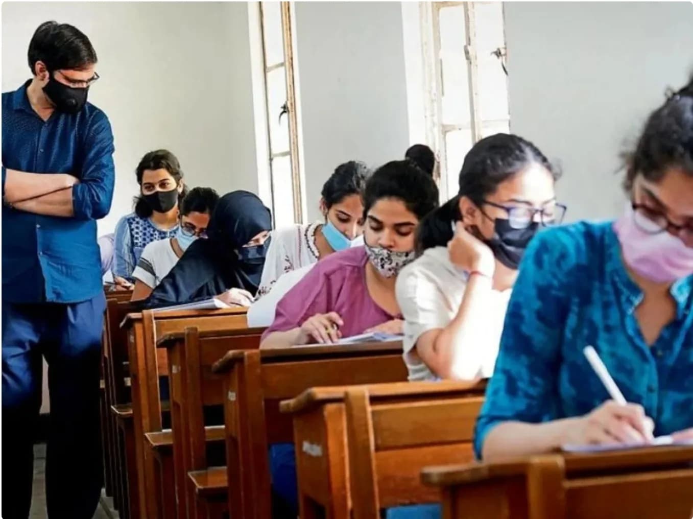 एक परीक्षा हॉल में छात्र परीक्षा दे रहे हैं और एक शिक्षक उन पर नजर रख रहा है।