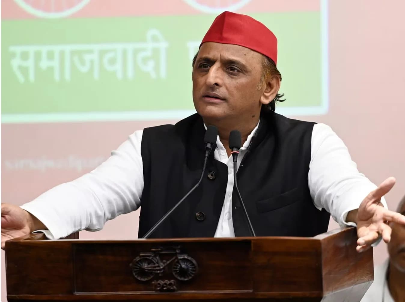 एक व्यक्ति लाल टोपी और काले कोट में पोडियम पर बोल रहा है।