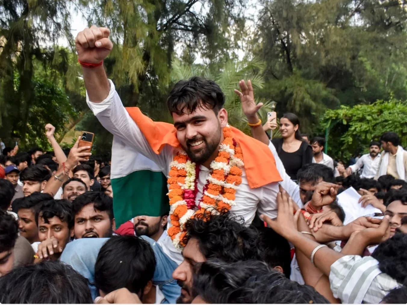 एक आदमी को भीड़ में कंधों पर उठाया गया है, जिसने नारंगी और सफेद फूलों की माला पहनी है।