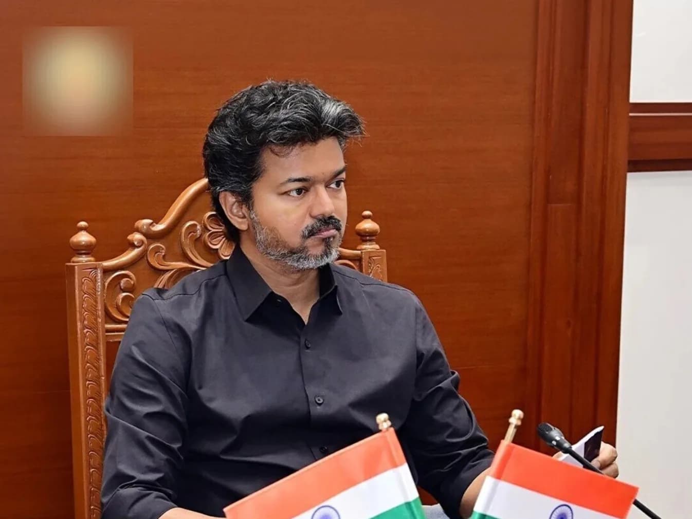 एक व्यक्ति काले रंग की शर्ट पहने हुए कुर्सी पर बैठा है, उसके सामने दो छोटे भारतीय झंडे हैं।