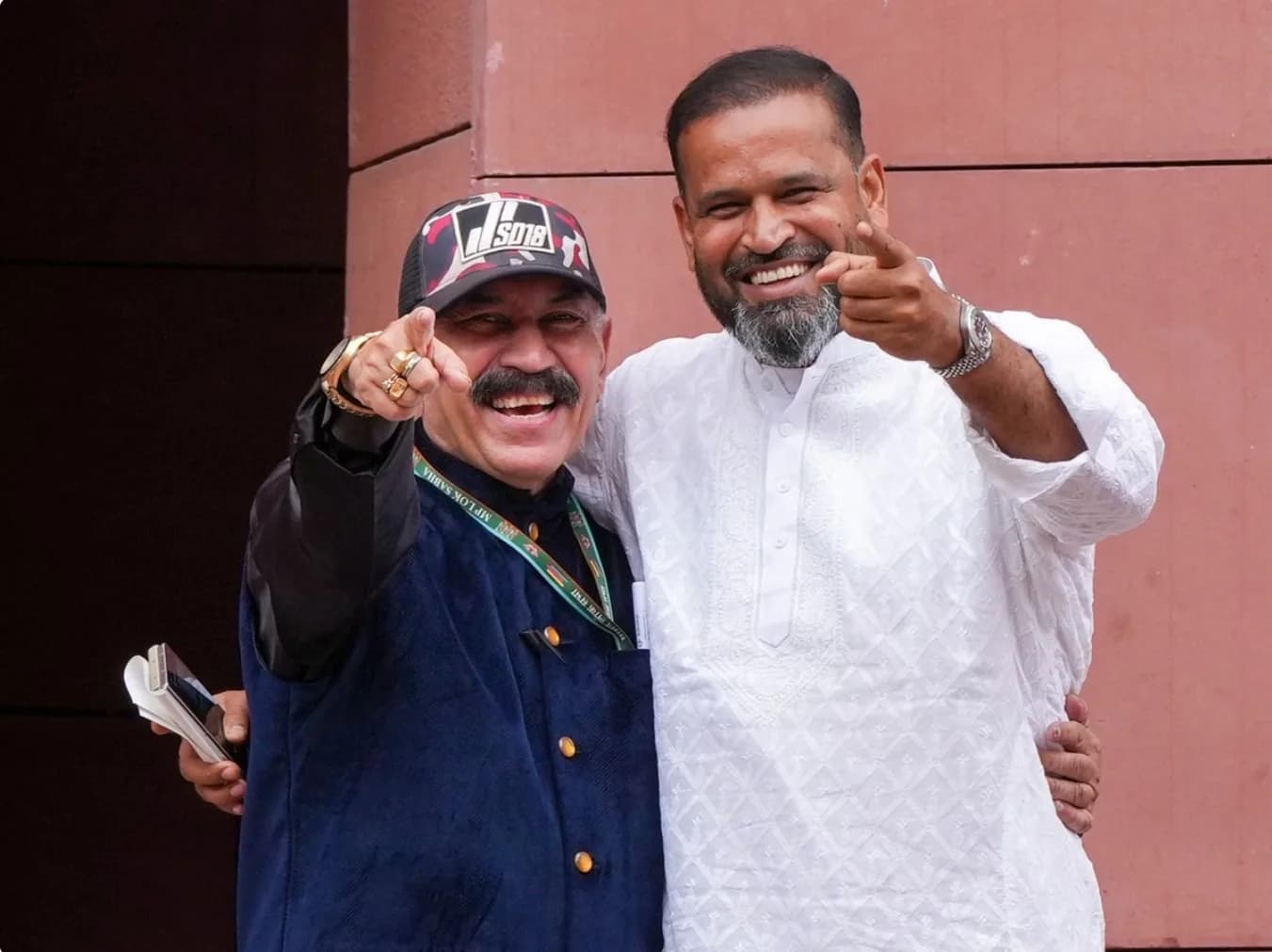 दो आदमी कैमरे की ओर इशारा करते हुए मुस्कुरा रहे हैं।