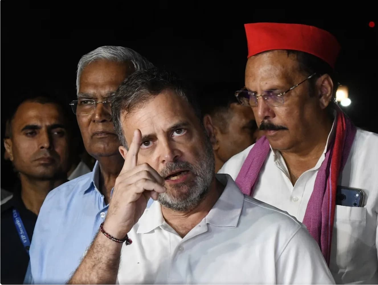 राहुल गांधी एक सफेद पोलो शर्ट पहने हुए हैं, उनके पीछे दो अन्य पुरुष खड़े हैं।