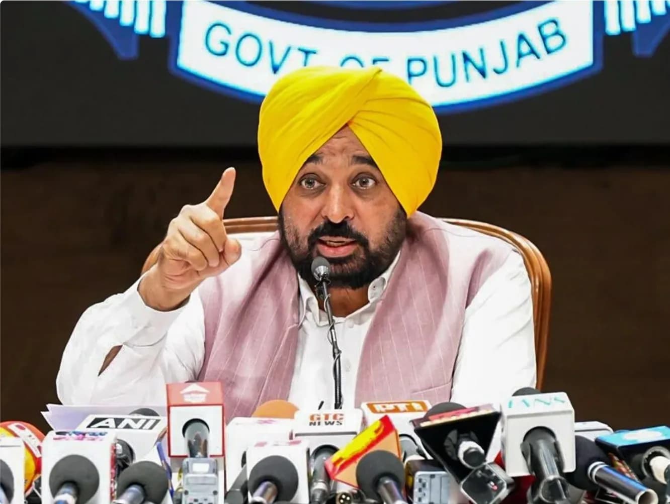 एक व्यक्ति पीले रंग की पगड़ी पहने हुए, माइक्रोफोन के सामने बोल रहा है और उंगली उठा रहा है।