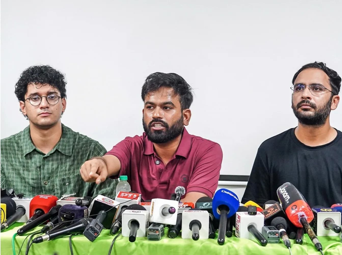 तीन आदमी एक प्रेस कॉन्फ्रेंस में बैठे हैं, बीच वाला आदमी बोल रहा है और उंगली उठा रहा है।