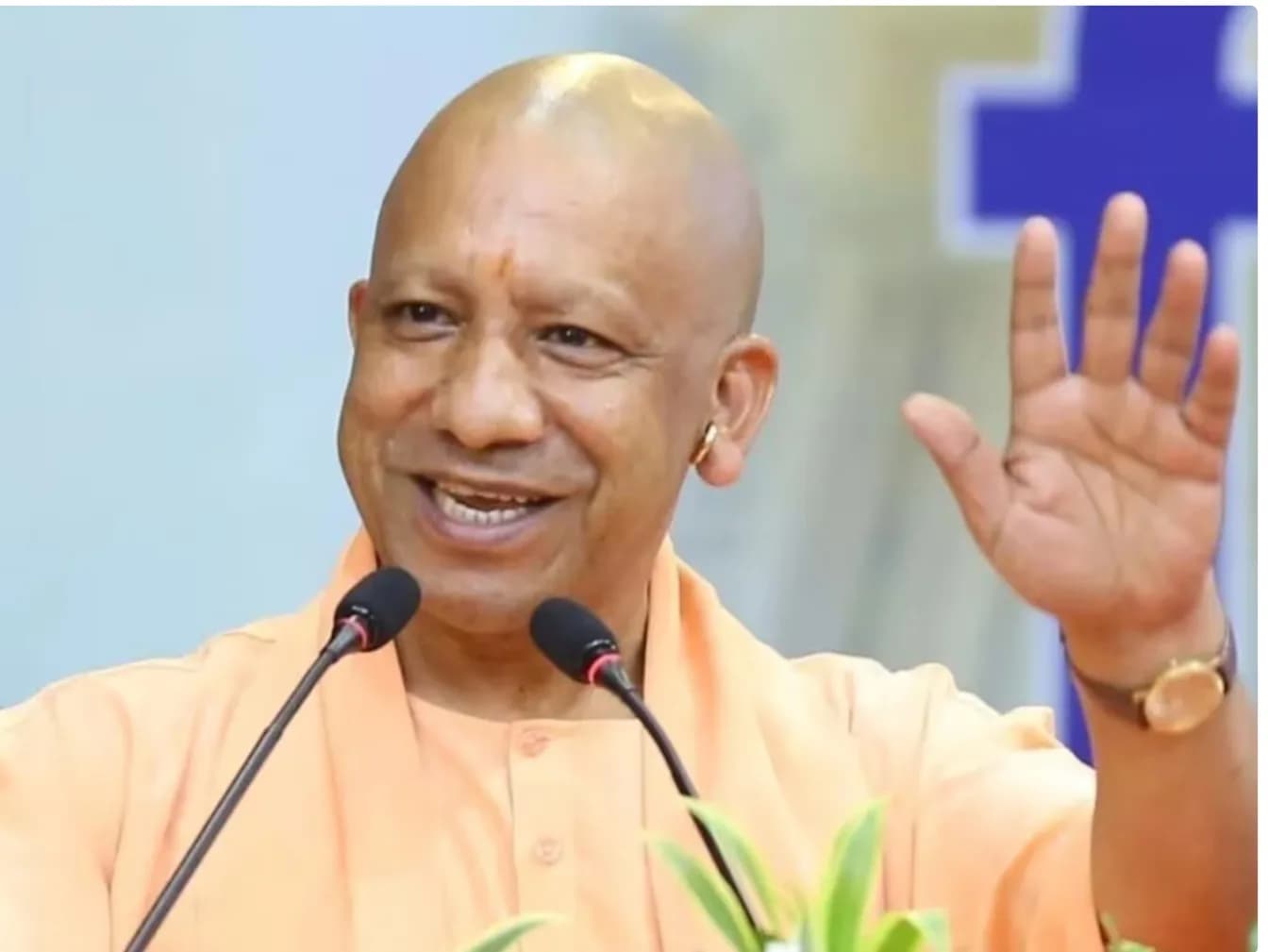 एक व्यक्ति नारंगी रंग के कपड़े पहने हुए, मुस्कुराते हुए और अपना हाथ ऊपर उठाए हुए है।