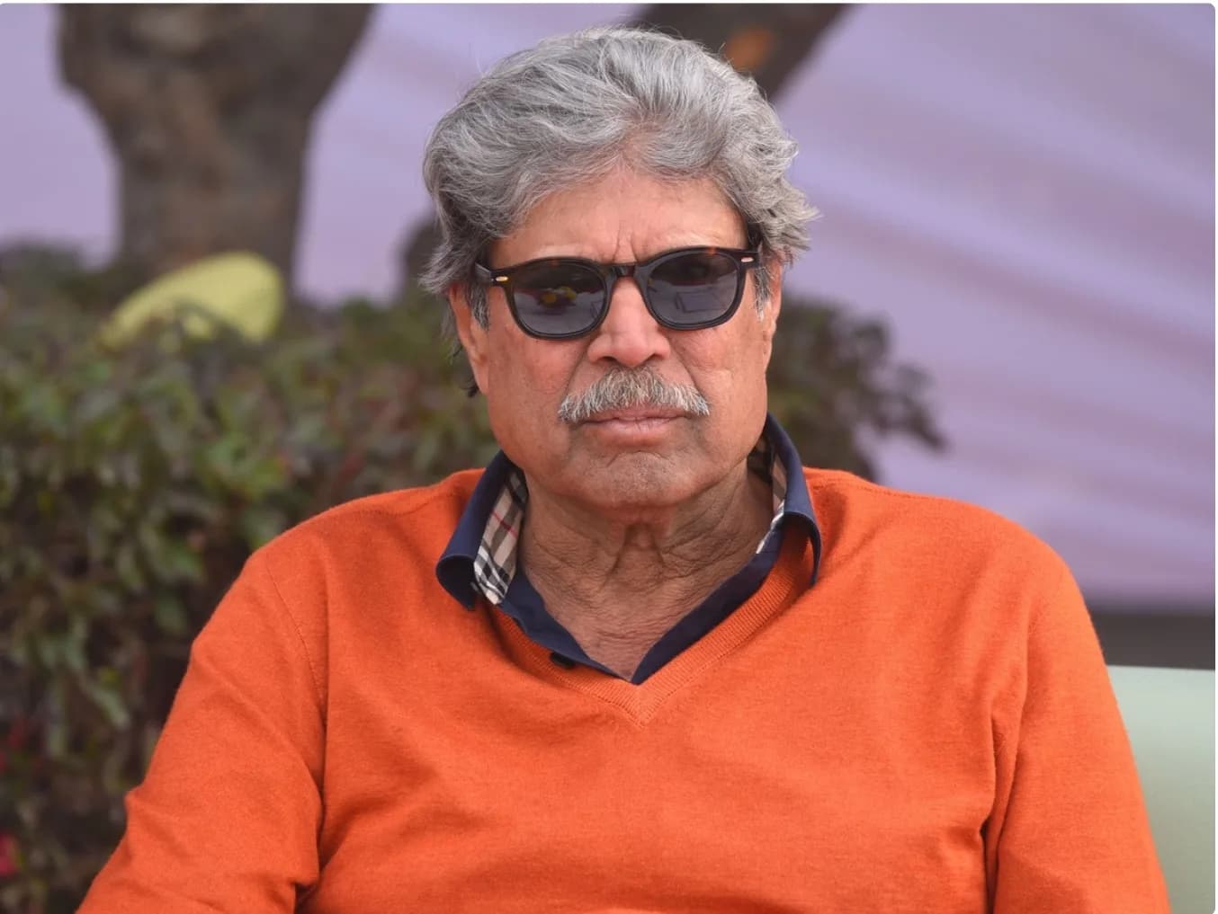 एक आदमी धूप का चश्मा और नारंगी स्वेटर पहने हुए है।