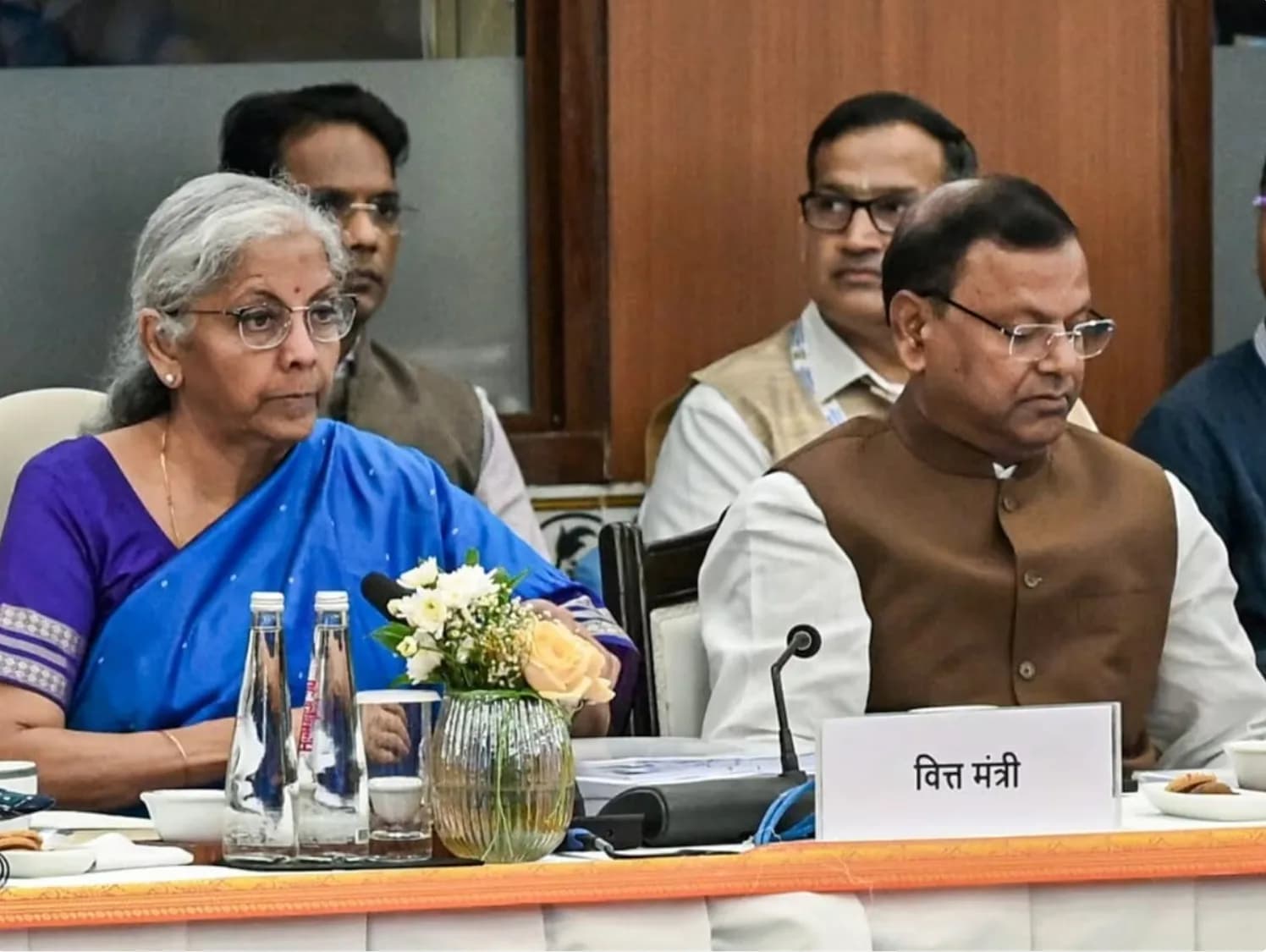 वित्त मंत्री एक मेज पर बैठे हैं, उनके सामने पानी की बोतलें और फूल हैं।
