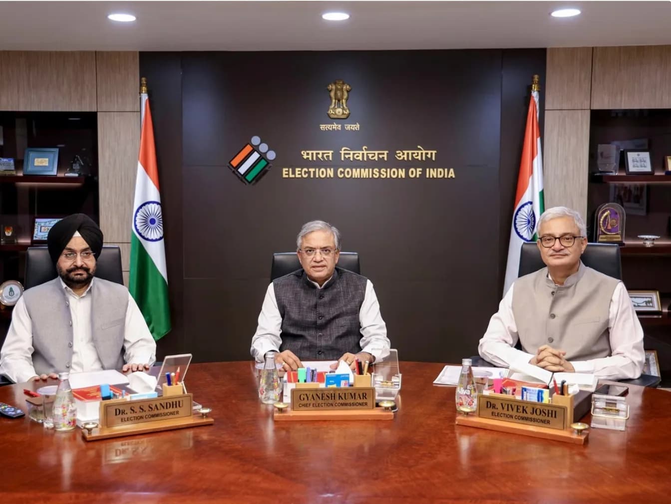 तीन अधिकारी भारत के चुनाव आयोग के लोगो के सामने बैठे हैं।