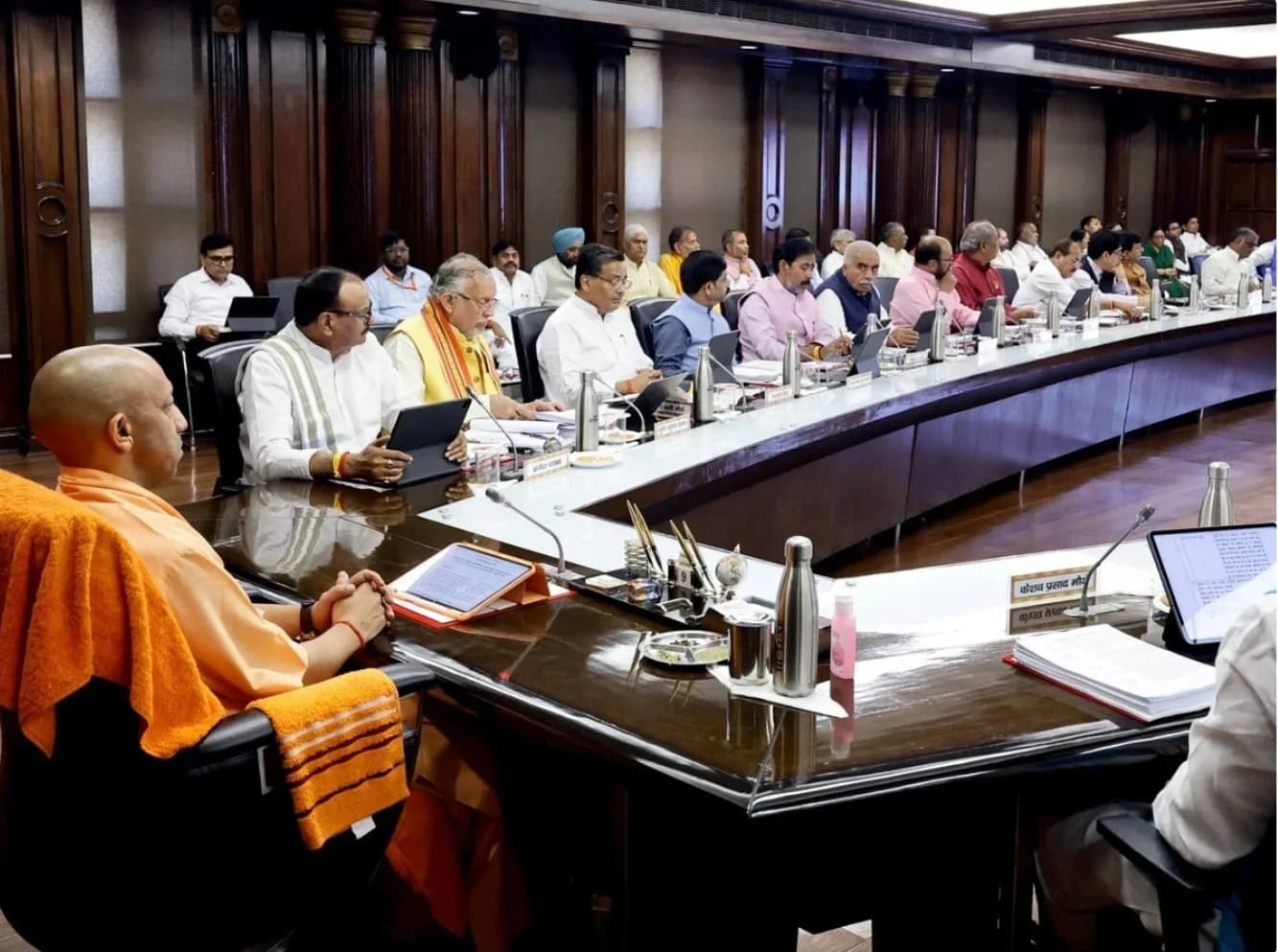 एक बड़े सम्मेलन कक्ष में एक लंबी मेज पर बैठे कई लोग एक बैठक में भाग ले रहे हैं।