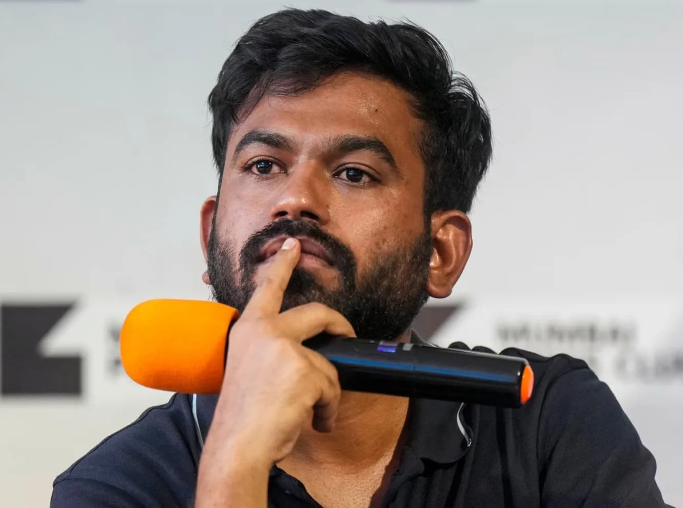 एक आदमी माइक्रोफ़ोन पकड़े हुए और सोचता हुआ दिखाई दे रहा है।