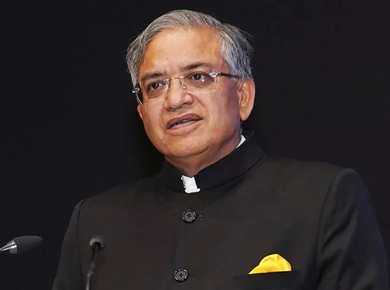 एक व्यक्ति काले रंग के कोट और सफेद शर्ट में, चश्मा पहने हुए, बोल रहा है।