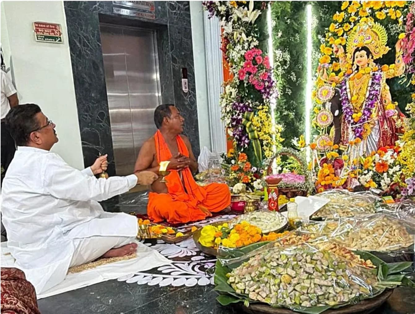 दो लोग एक देवी की मूर्ति के सामने फर्श पर बैठे हैं, जिसके चारों ओर फूल और प्रसाद है।