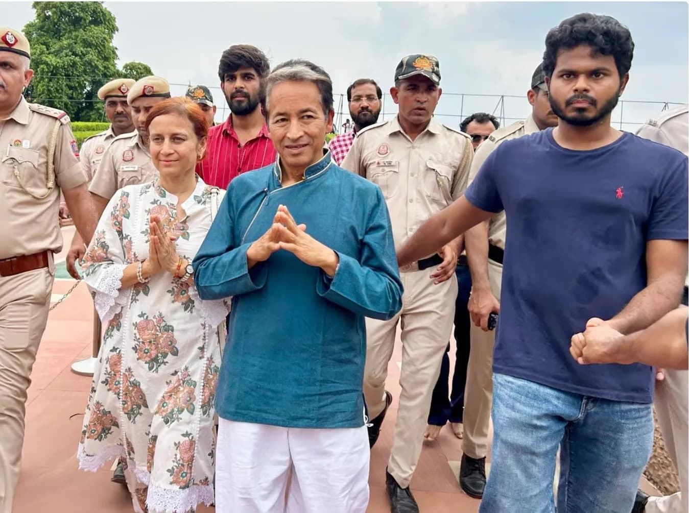 एक महिला और एक पुरुष नमस्ते की मुद्रा में खड़े हैं, उनके आसपास कई लोग हैं।