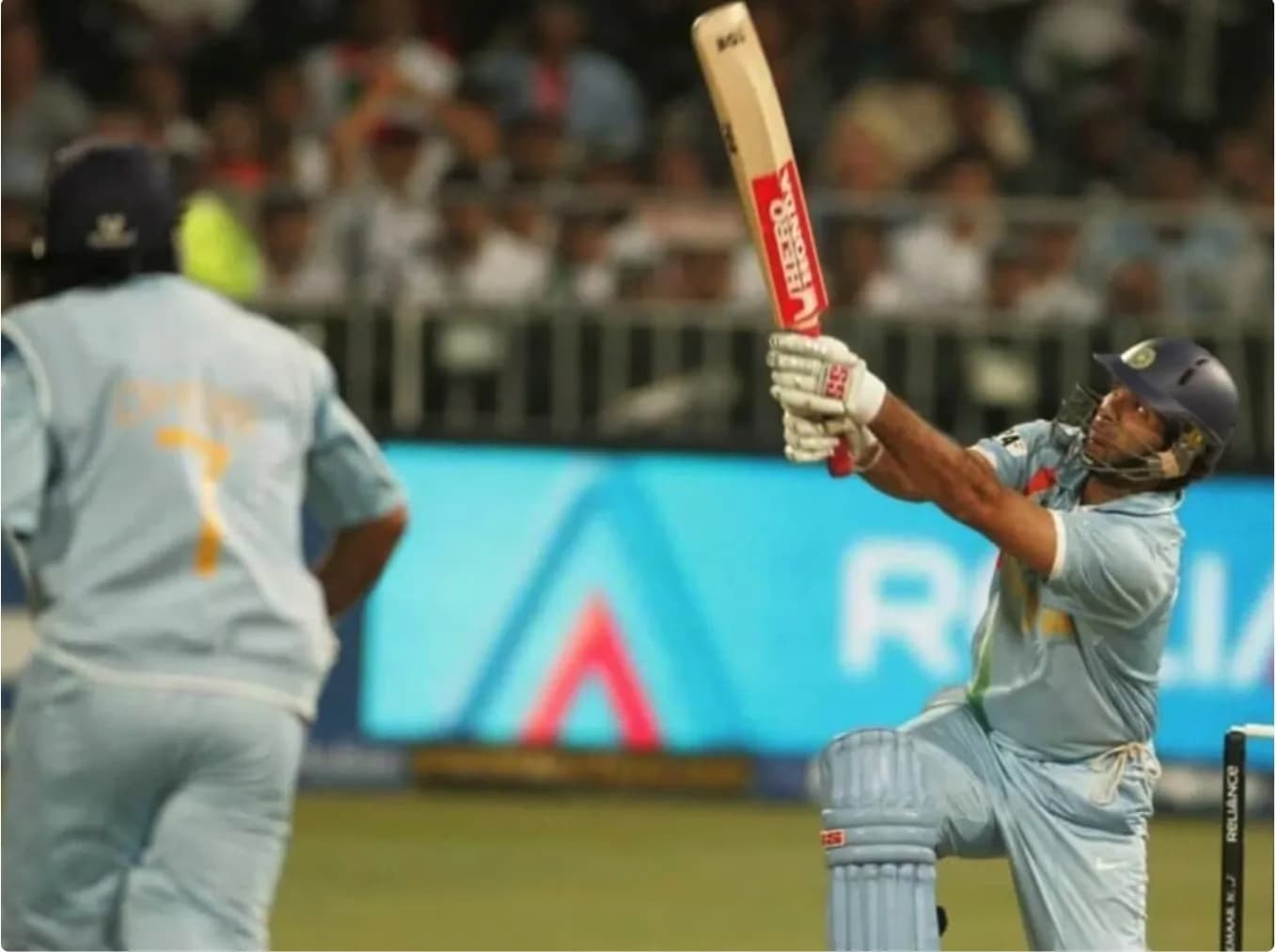 एक क्रिकेट खिलाड़ी नीली जर्सी में क्रिकेट बैट पकड़े हुए है, जबकि दूसरा खिलाड़ी उसके पीछे खड़ा है।