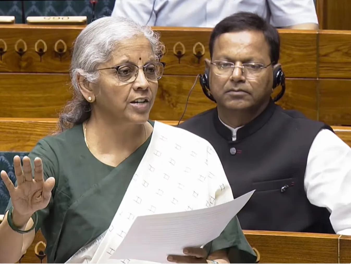एक महिला हरे रंग की साड़ी और सफेद शॉल में बोल रही है, उसके पीछे एक आदमी हेडफ़ोन पहने बैठा है।