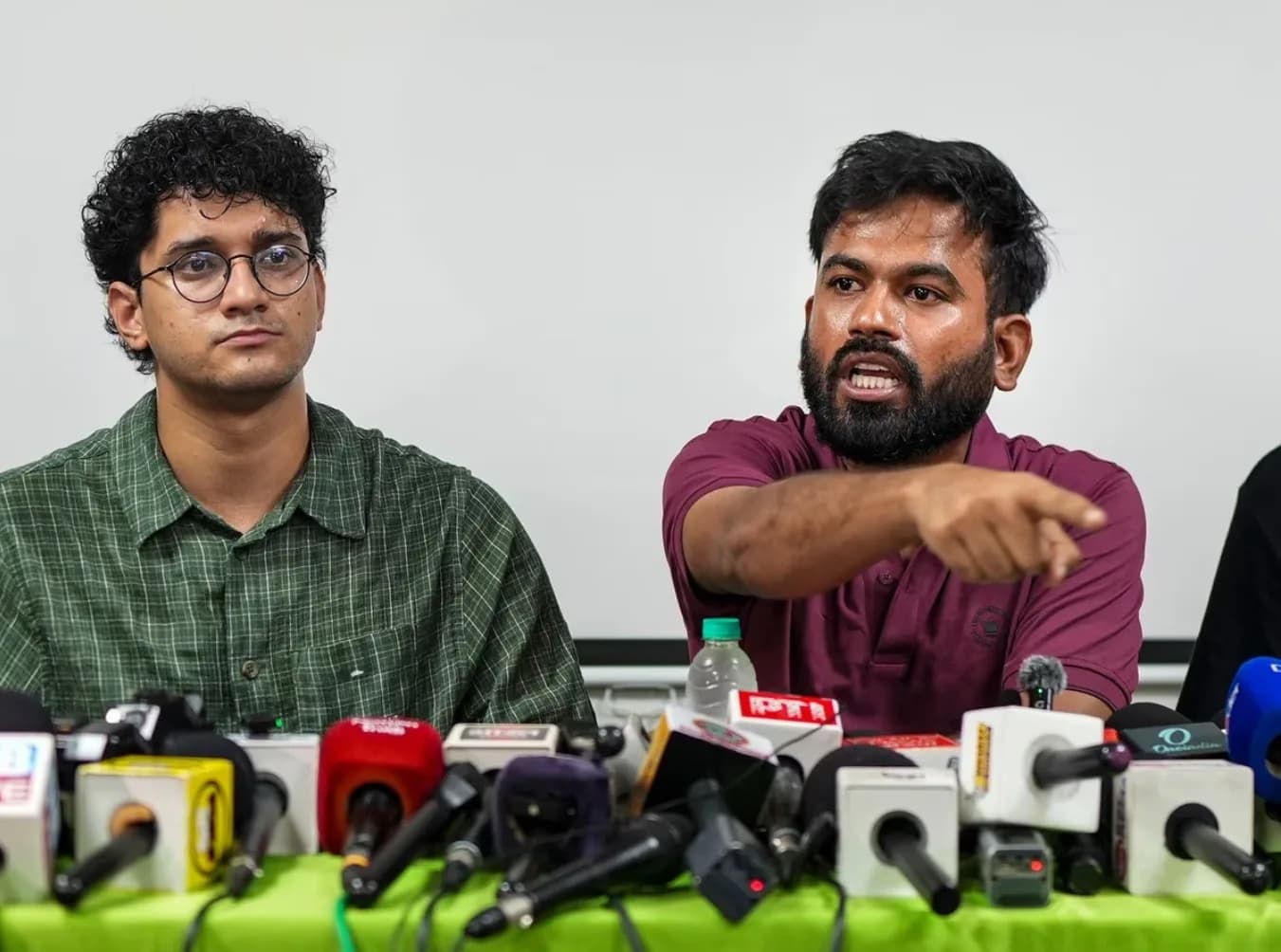 माइक्रोफोन से घिरी मेज पर दो पुरुष बैठे हैं, एक इशारा कर रहा है।