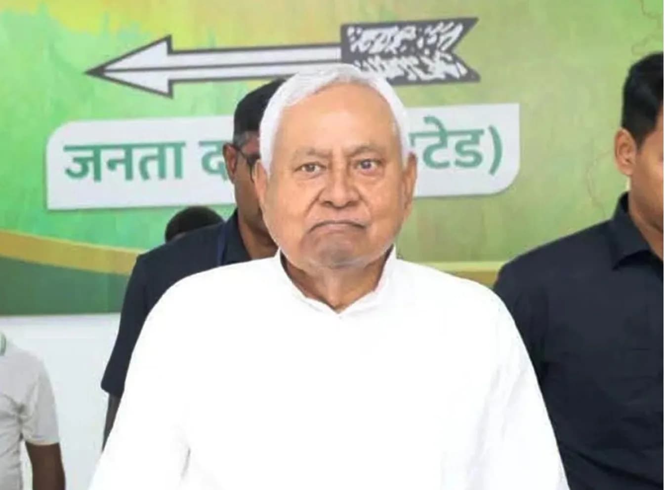 एक व्यक्ति सफेद कुर्ता पहने हुए है, जिसके पीछे एक बैनर है।