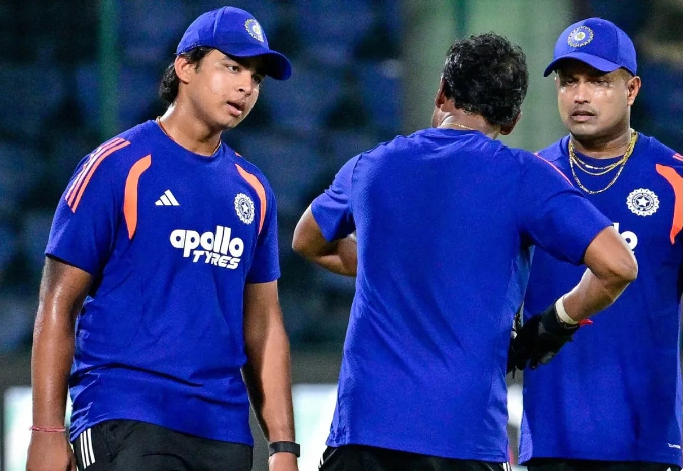 तीन क्रिकेट खिलाड़ी नीली जर्सी और टोपी पहने हुए मैदान पर बातचीत कर रहे हैं।