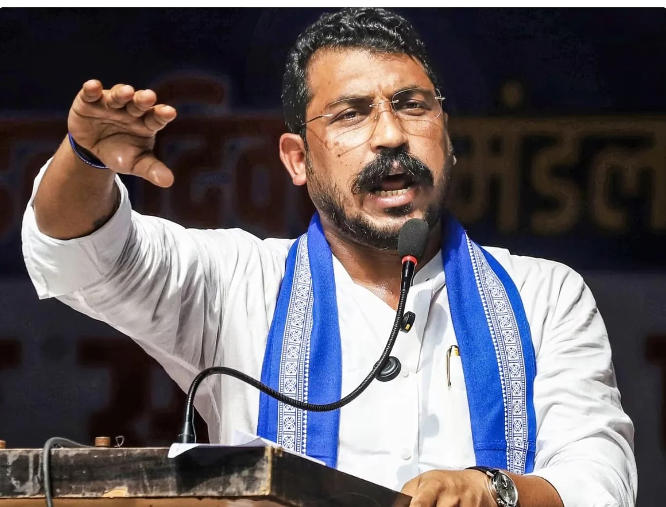 एक व्यक्ति सफेद शर्ट और नीले दुपट्टे में माइक पर बोल रहा है, उसका एक हाथ उठा हुआ है।